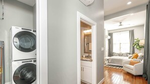 Una sala de bugaderia amb dues piles de rentadores i assecadores frontals, una cuina americana petita amb taulell i armaris, i una zona d'estar amb un sofà i una taula de centre.