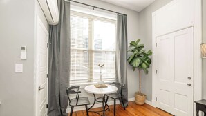 A room with a wooden floor, a white door, a window with grey curtains, a round table with two chairs, a potted plant, and a thermostat on the wall. - Findlay Market Suite 1 – For 2! (Cincinnati)
