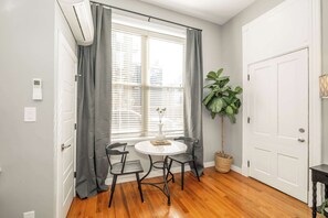 A room with a wooden floor, a white door, a window with grey curtains, a round table with two chairs, a potted plant, and a thermostat on the wall. - Findlay Market Suite 1 – For 2! (Cincinnati)