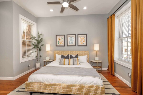 A bedroom with a bed, two bedside tables, a ceiling fan, and three framed artworks on the wall.