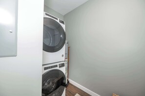 A laundry room with a stacked washer and dryer, a broom, and a small rug on the floor.