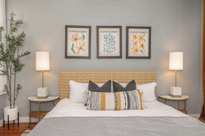 A neatly arranged bedroom with a large bed, two bedside tables with lamps, a potted plant, and three framed floral artwork on the wall.