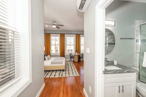 A hotel room with a bed, a desk, a chair, a round table, a bathroom with a sink and mirror, and a shower.