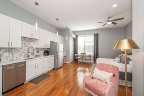Une cuisine moderne avec des armoires blanches, un pare-feu en marbre et un lave-vaisselle. On y trouve un coin repas avec une table et des chaises, ainsi qu’une chambre à coucher avec un lit et un fauteuil rose.