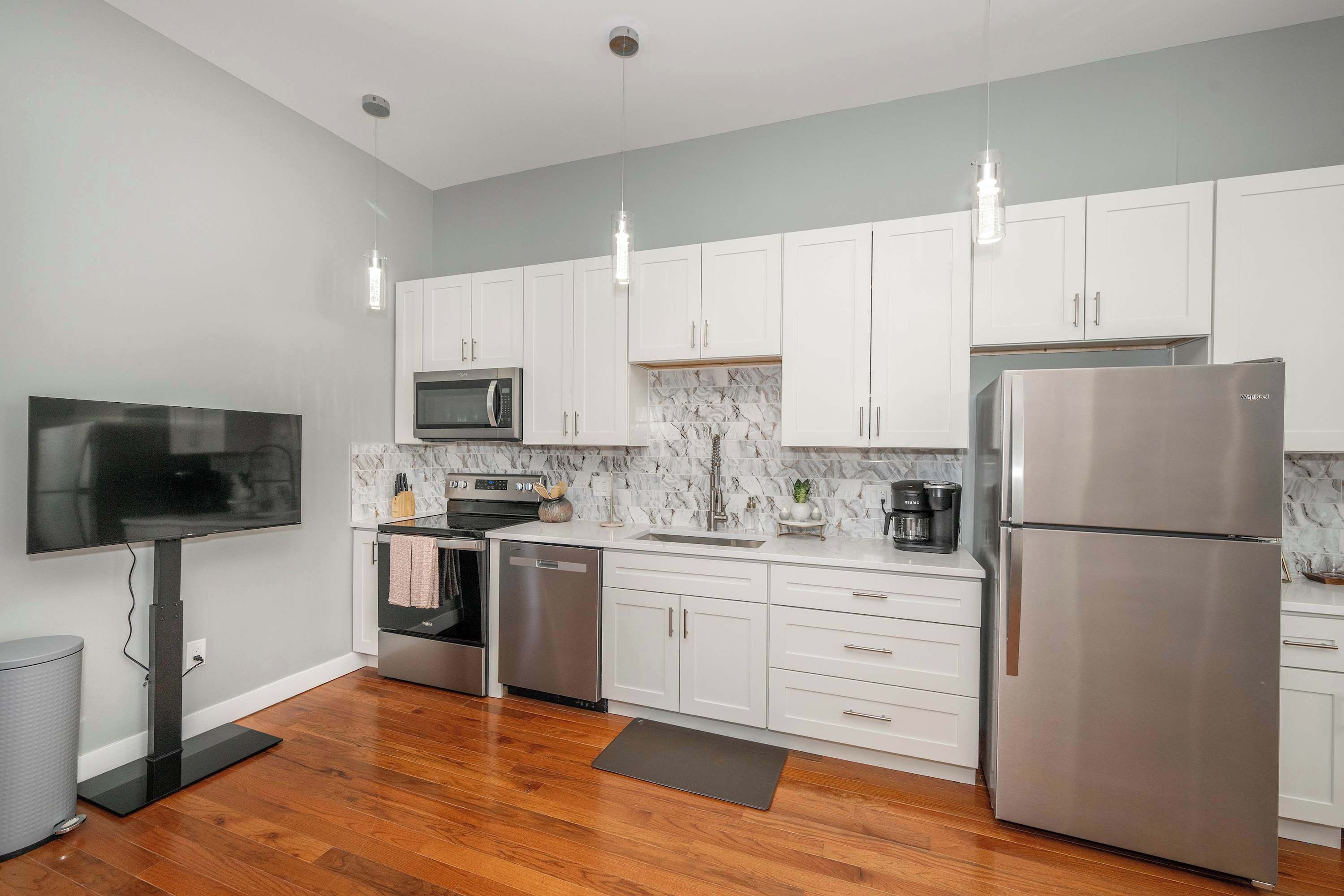 A modern kitchen with white cabinets, stainless steel appliances, a flat-screen TV, and wooden flooring.