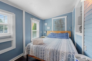 A bedroom with a bed, two windows with blinds, a bedside table, a lamp, and a wall-mounted shelf.