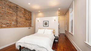 Une chambre avec un mur en briques, un lit recouvert de linge de lit blanc, un plancher en bois et une porte donnant sur une autre pièce.