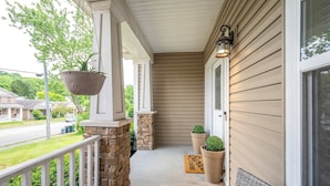 A covered porch with a stone column, potted plants, a doormat, and a hanging planter. - Urban Escape (Cincinnati)