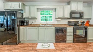 A kitchen with light green cabinets, stainless steel appliances, and a window with a view of greenery. - Dragonfly Cove | Private Dock! (Mooresville)