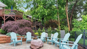 Een terras met Adirondack-stoelen en een overdekte tafel, omgeven door weelderige begroeiing en een houten terras.