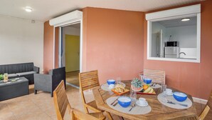 A dining area with a wooden table set for four, surrounded by wicker chairs. A kitchen is visible through an open doorway. - Apartment Alizés - Sea view & Direct beach access (Le quartier le Coin)
