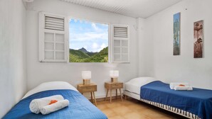 A room with two beds, a window with a view of green hills, and two bedside tables with lamps. - Apartment Alizés - Sea view & Direct beach access (Le quartier le Coin)