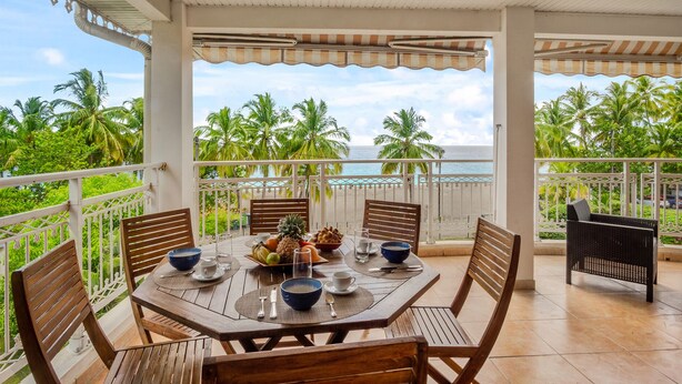 A wooden table set for dining with a view of the ocean and palm trees. - Apartment Alizés - Sea view & Direct beach access (Le quartier le Coin)