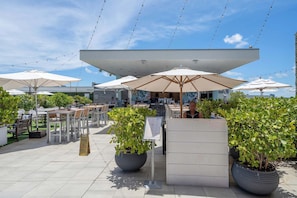 An outdoor seating area with tables and umbrellas, surrounded by potted plants and a clear sky.