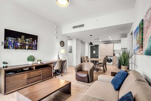 Ein modernes Wohnzimmer mit Couchtisch aus Holz, einem beigefarbenen Sofa mit blauen Kissen, einem an der Wand montierten Fernseher und einem Stadtbildgemälde.