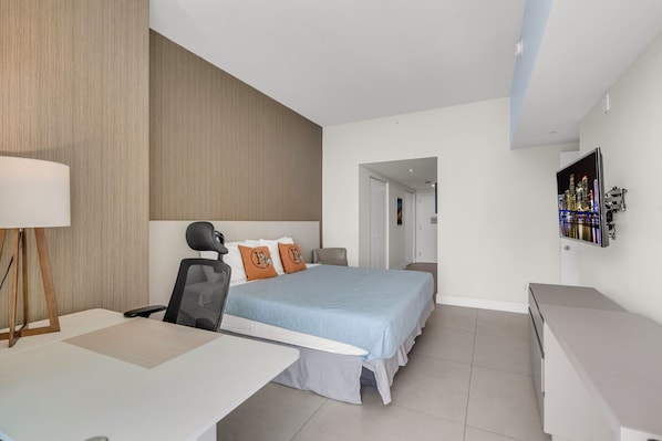 A modern hotel room with a bed, desk, and chair, a TV on the wall, and a door leading to another room.