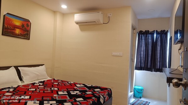 A bedroom with a bed, a wall-mounted air conditioner, a painting, and a bathroom with a shower curtain and a blue trash bin. - YADUKUL HOTEL (Hyderabad)