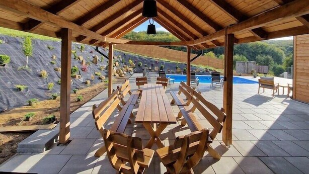 An outdoor patio with wooden benches and a table, surrounded by a swimming pool and a landscaped garden. - Holiday Home Wiśniowa with Pool and Mountain Views (Iwierzyce)