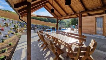 A covered outdoor seating area with wooden benches and a table, surrounded by a landscaped garden and a pool.