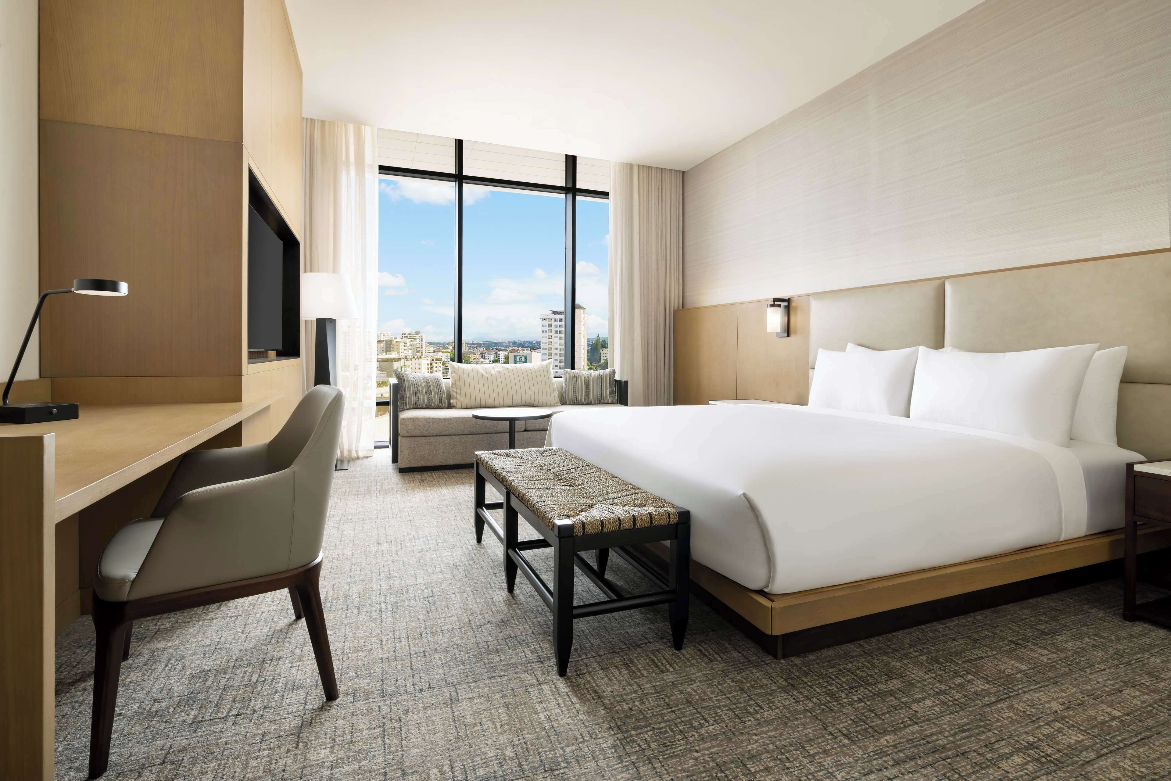 A hotel room with a large bed, a desk, a chair, a bench, and a TV.