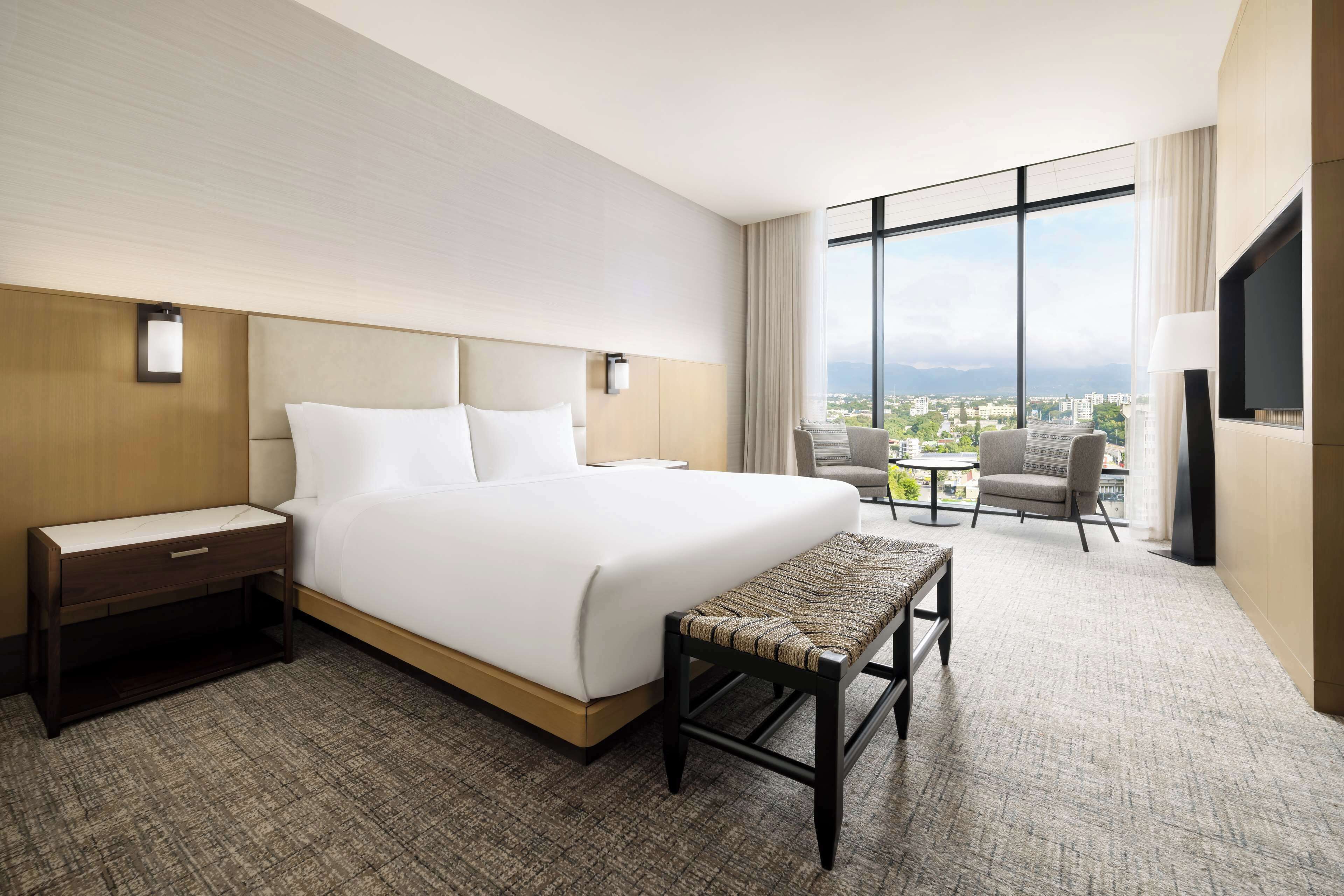 A hotel room with a large bed, a nightstand, a bench, a small table with chairs, and a large window offering a city view.