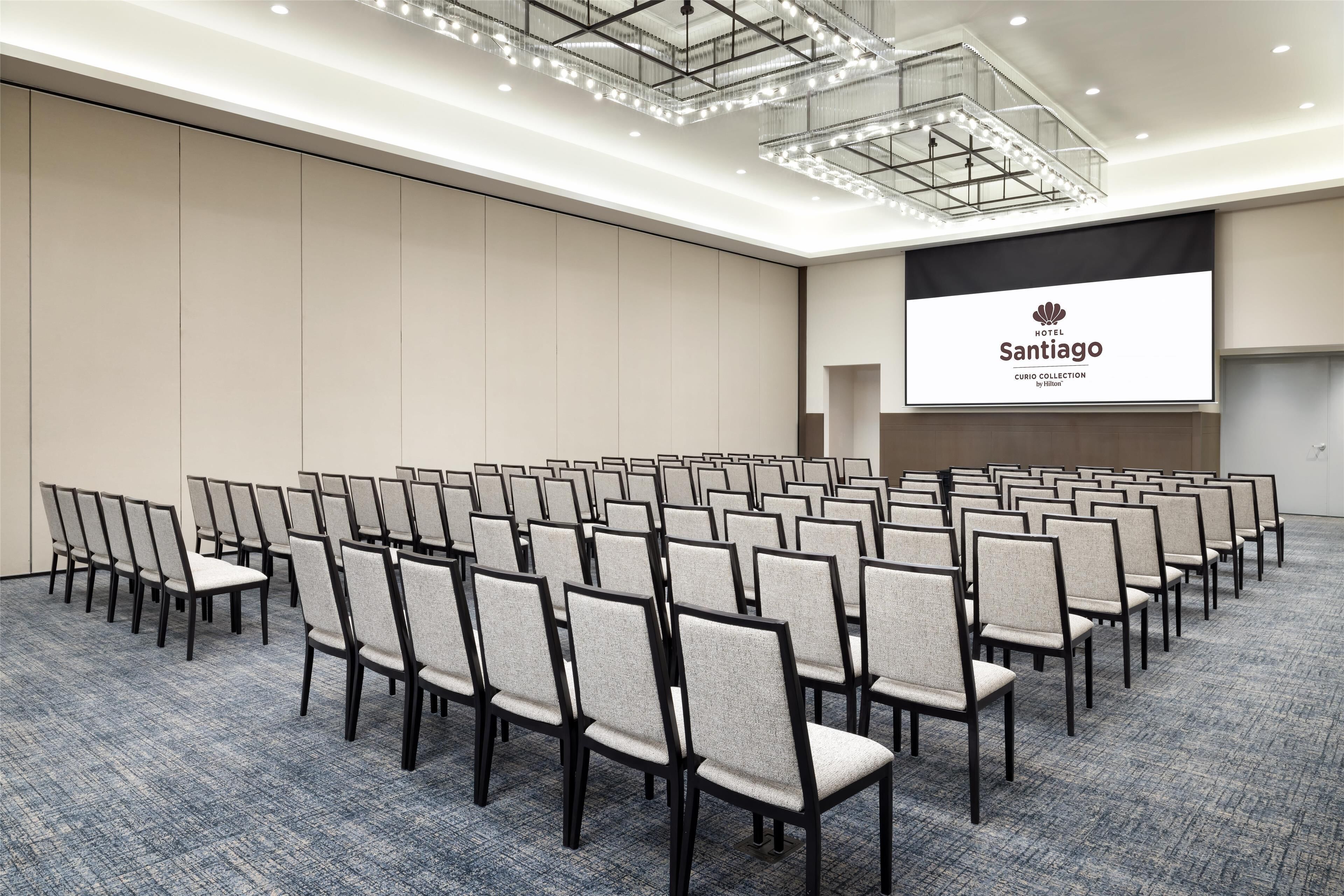 A conference room with rows of chairs, a large screen displaying the hotel name, and a modern chandelier.