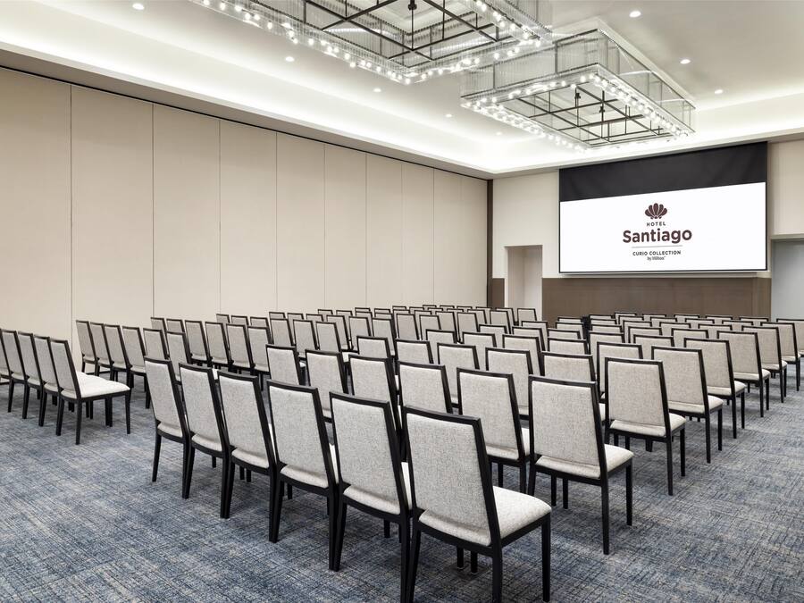 A conference room with rows of chairs, a large screen displaying the hotel name, and a modern chandelier.
