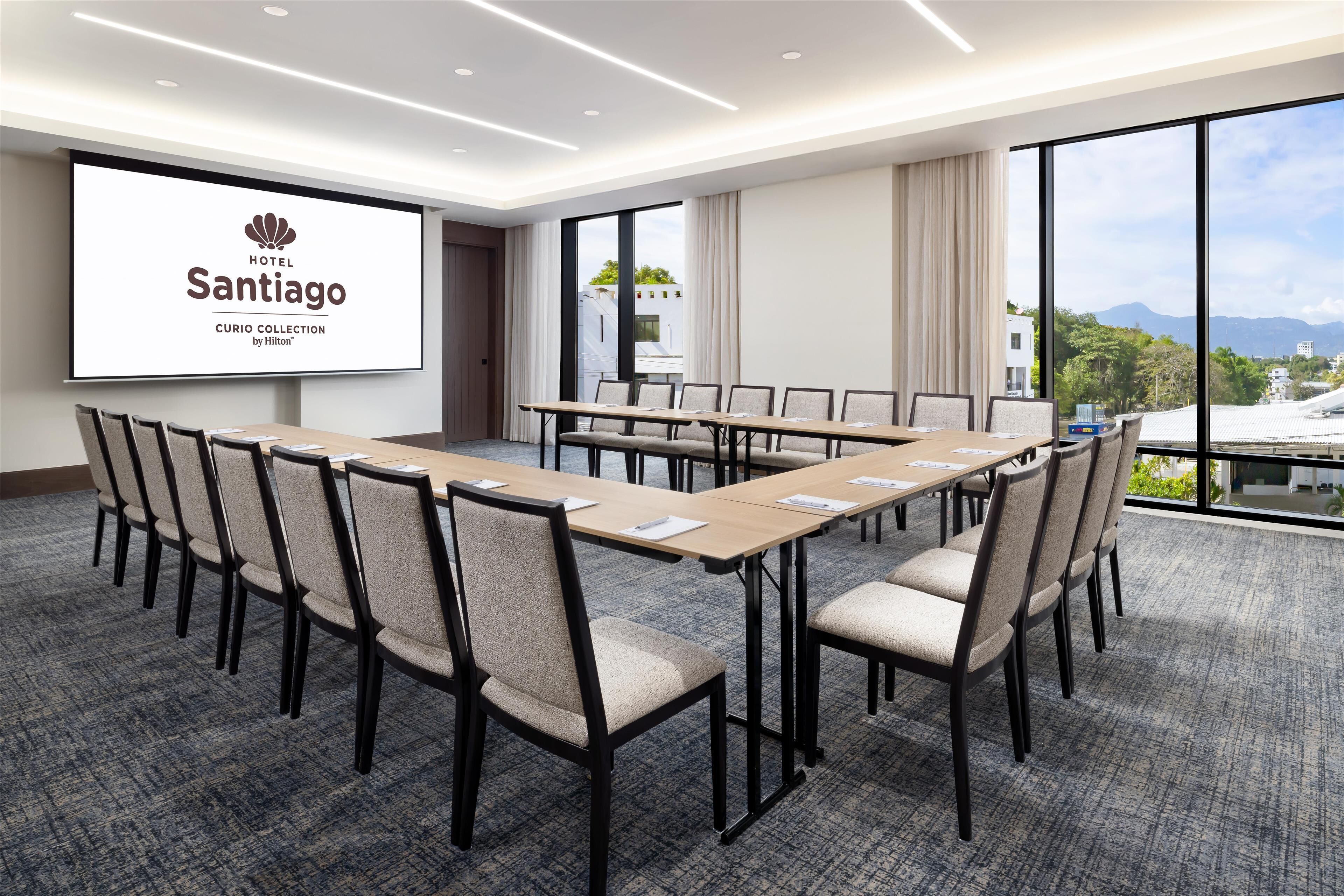 Ein Hotelkonferenzraum mit einer großen Leinwand, die den Namen des Hotels zeigt, einem langen Tisch mit Stühlen und einem Teppichboden.