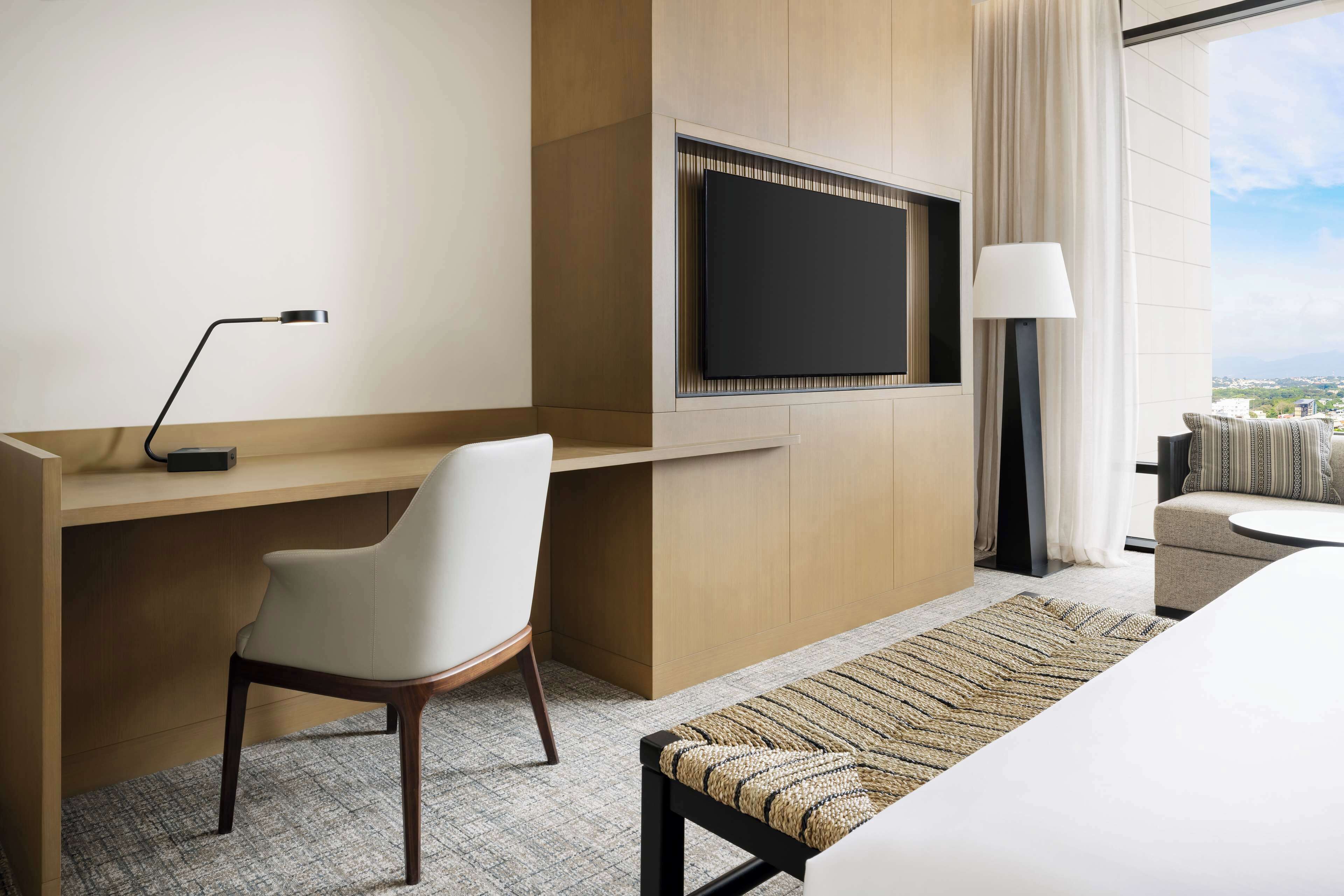 A modern hotel room with a desk, chair, TV, and a view of the city.