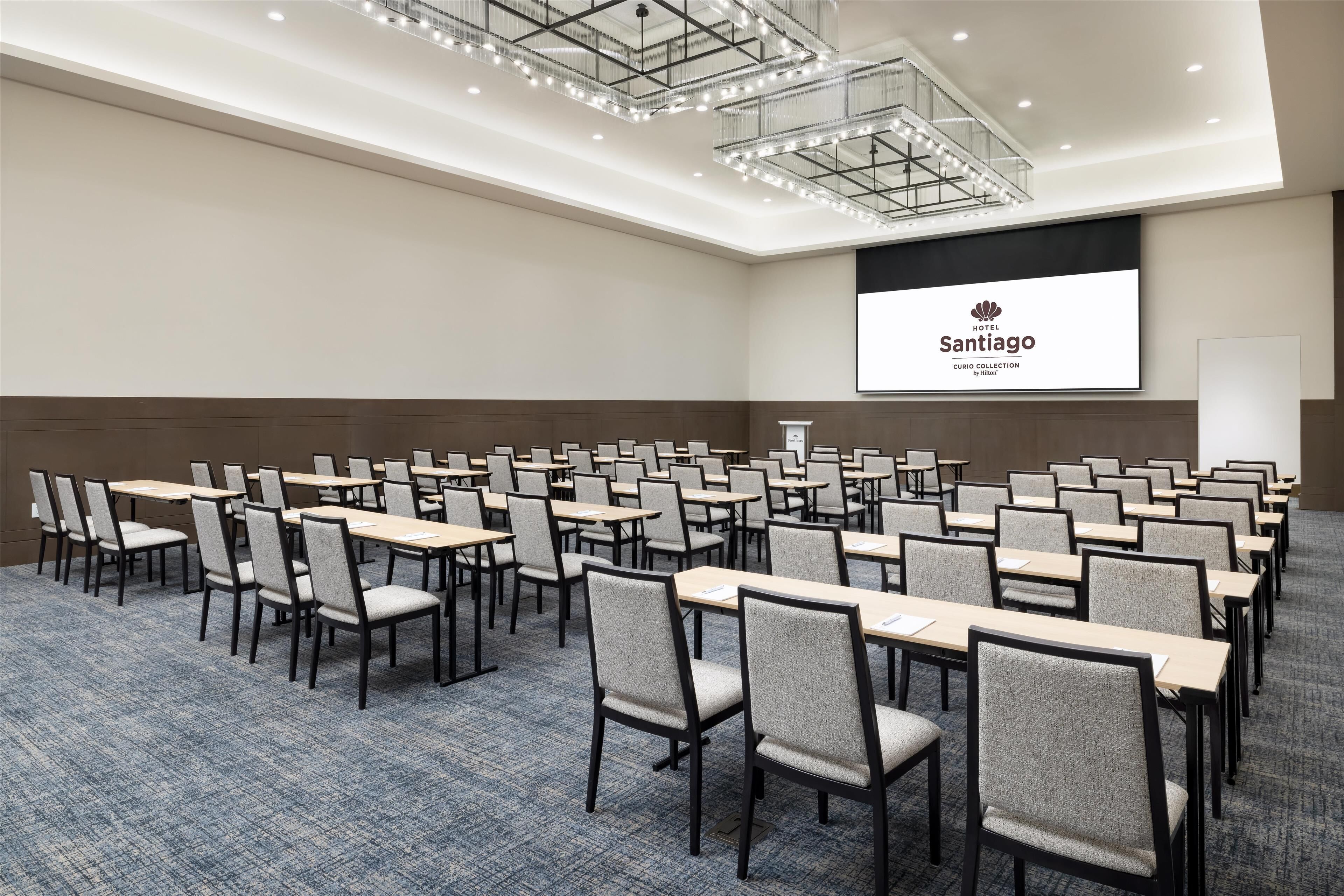 A conference room with rows of tables and chairs, a large screen displaying 'Hotel Santiago', and a modern ceiling design.