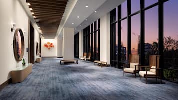 A spacious hallway with a modern design, featuring a long bench, seating area with chairs, and large windows offering a view of the cityscape at dusk.