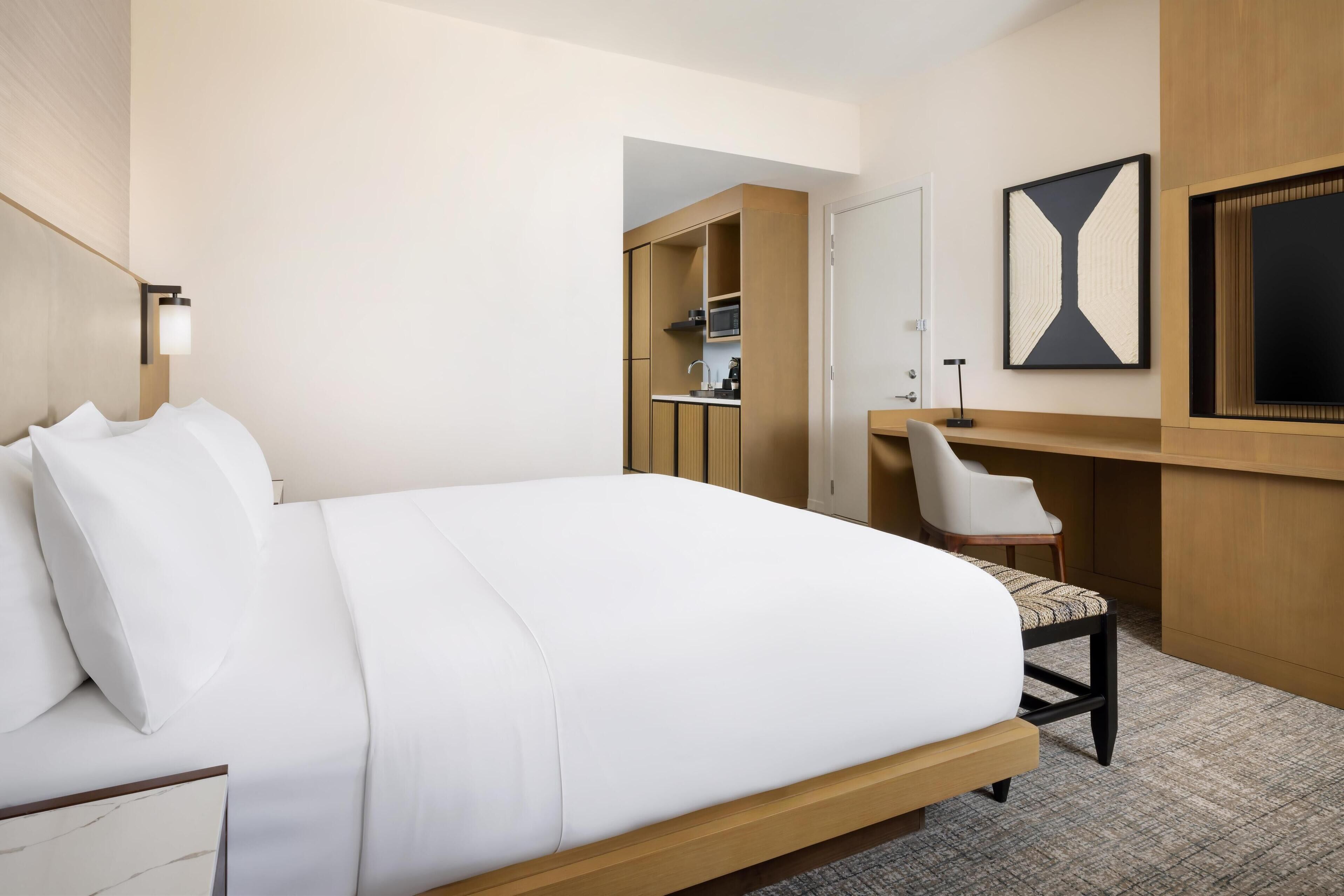 Ein Hotelzimmer mit einem großen Bett, einem Schreibtisch mit Stuhl, einer Küchenzeile und einem großen Fenster.