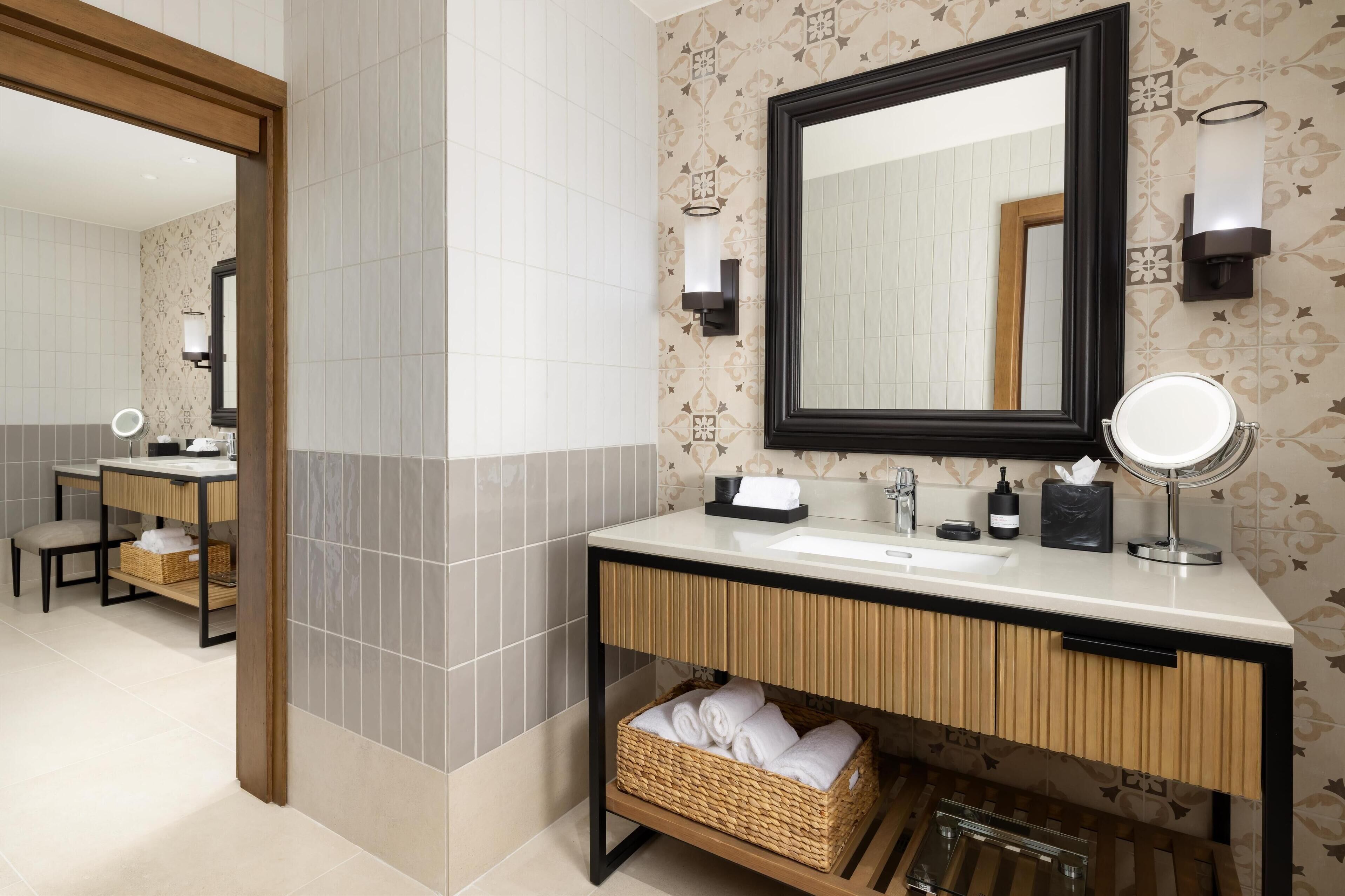 A bathroom with a large mirror, a sink, a vanity, and a wicker basket under the counter.