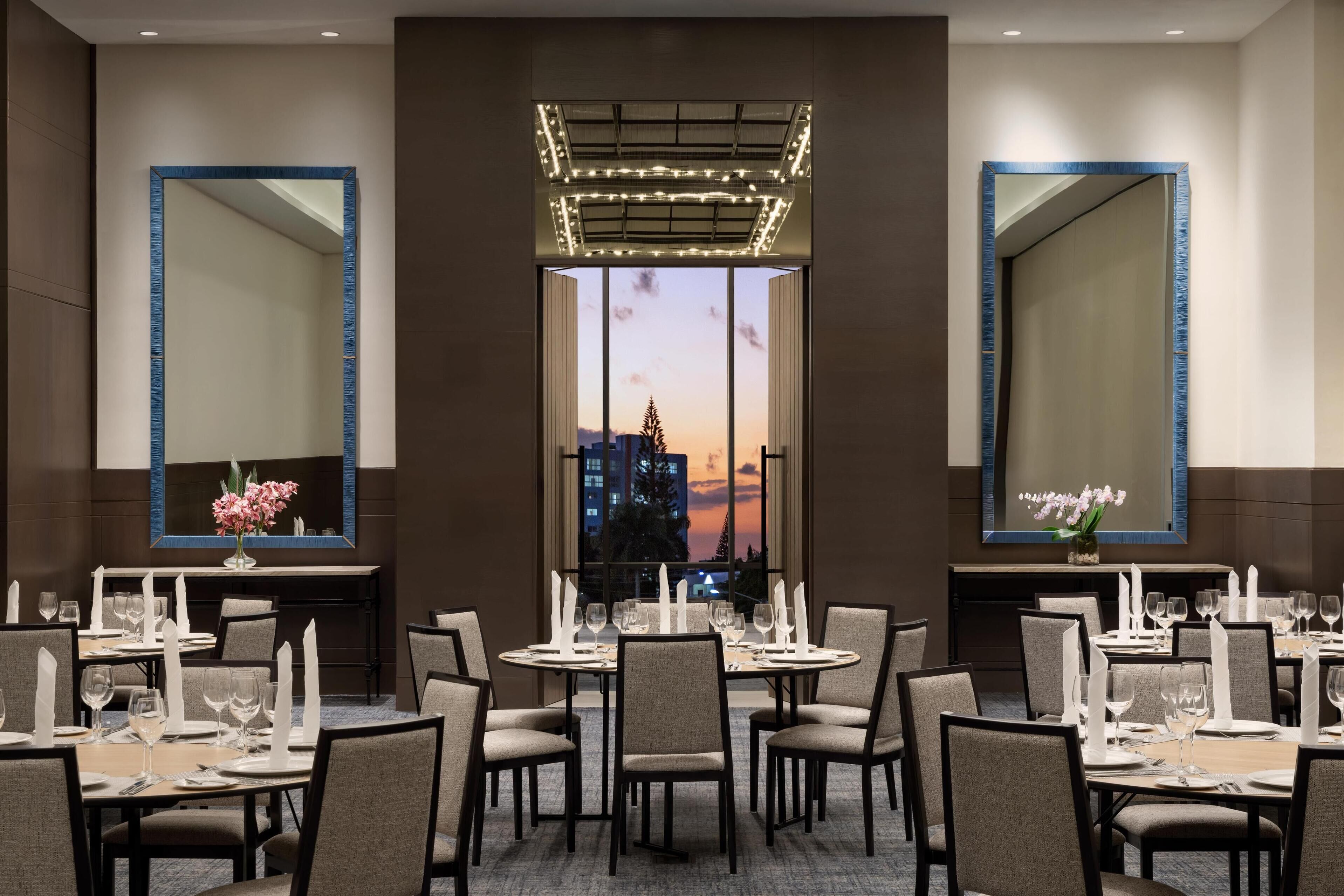 Un espace repas avec plusieurs tables dressées pour le dîner, de grands miroirs sur les murs et une vue sur le paysage urbain au coucher du soleil.