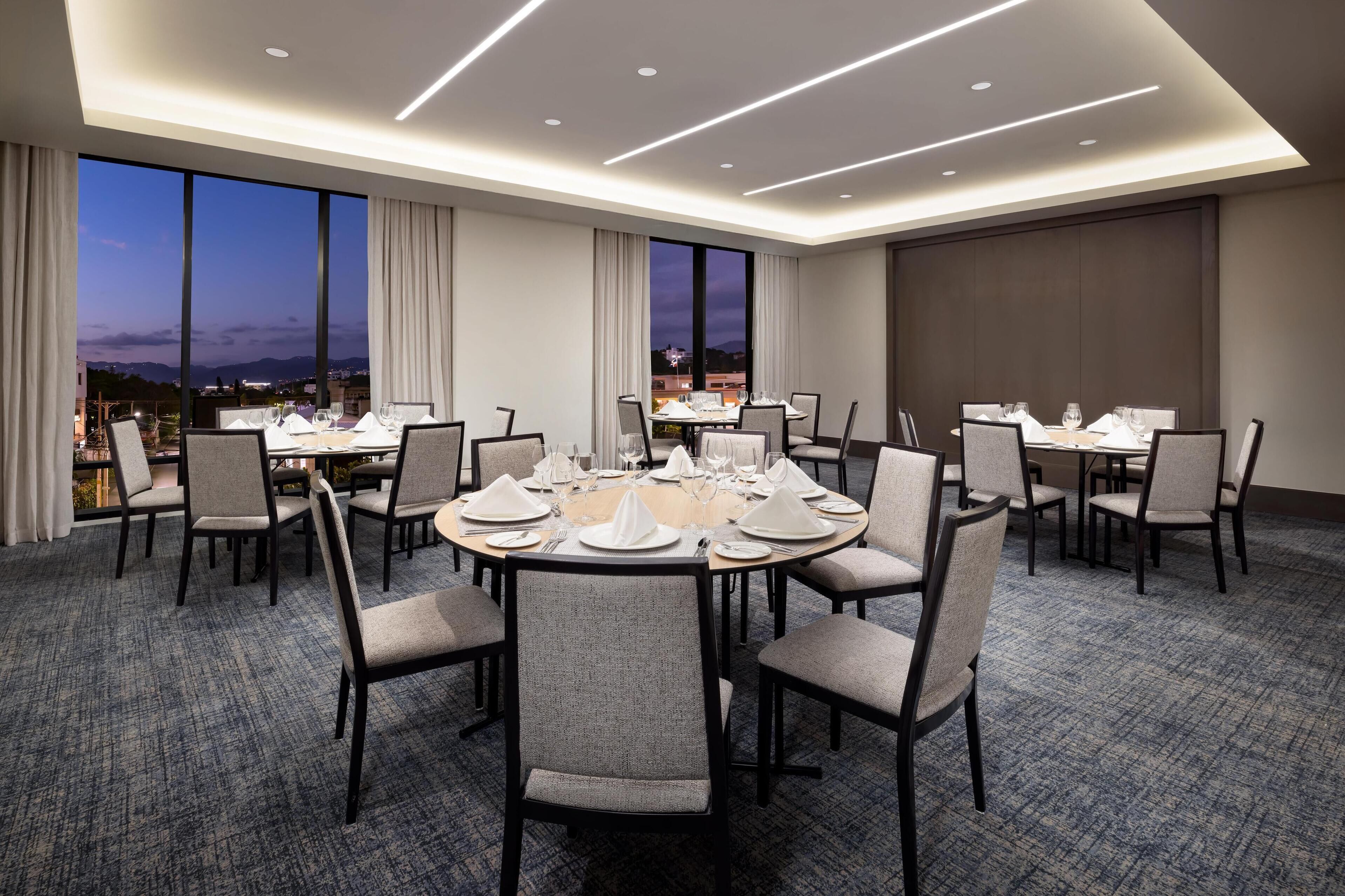 A conference room with round tables set for dining, large windows with curtains, and a carpeted floor.