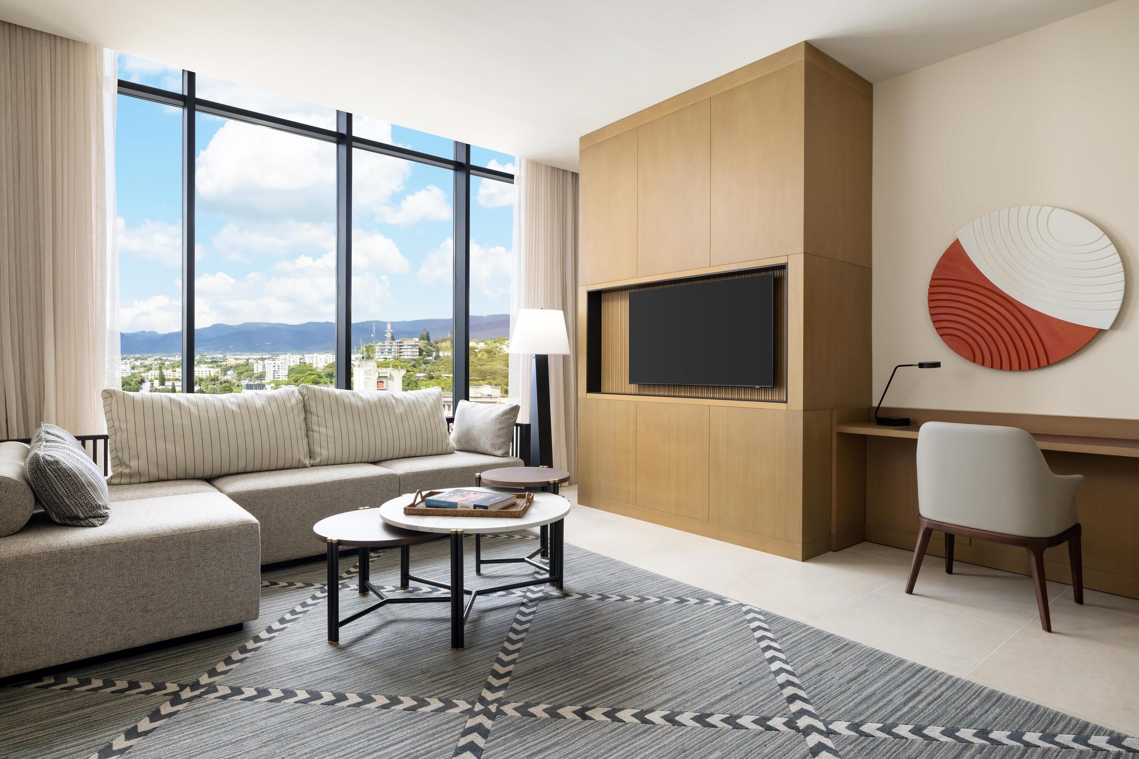 Habitación de hotel moderna con un ventanal grande que ofrece vistas a la ciudad, un sofá, una mesa de centro redonda, un escritorio y un televisor montado en la pared.