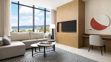 Habitación de hotel moderna con un ventanal grande que ofrece vistas a la ciudad, un sofá, una mesa de centro redonda, un escritorio y un televisor montado en la pared.