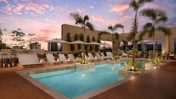 Zona de piscina con sillones de descanso y sombrillas, rodeada de palmeras y un cielo al atardecer.