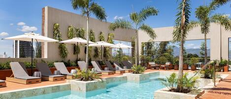 A poolside area with lounge chairs and umbrellas, surrounded by palm trees and a modern building.