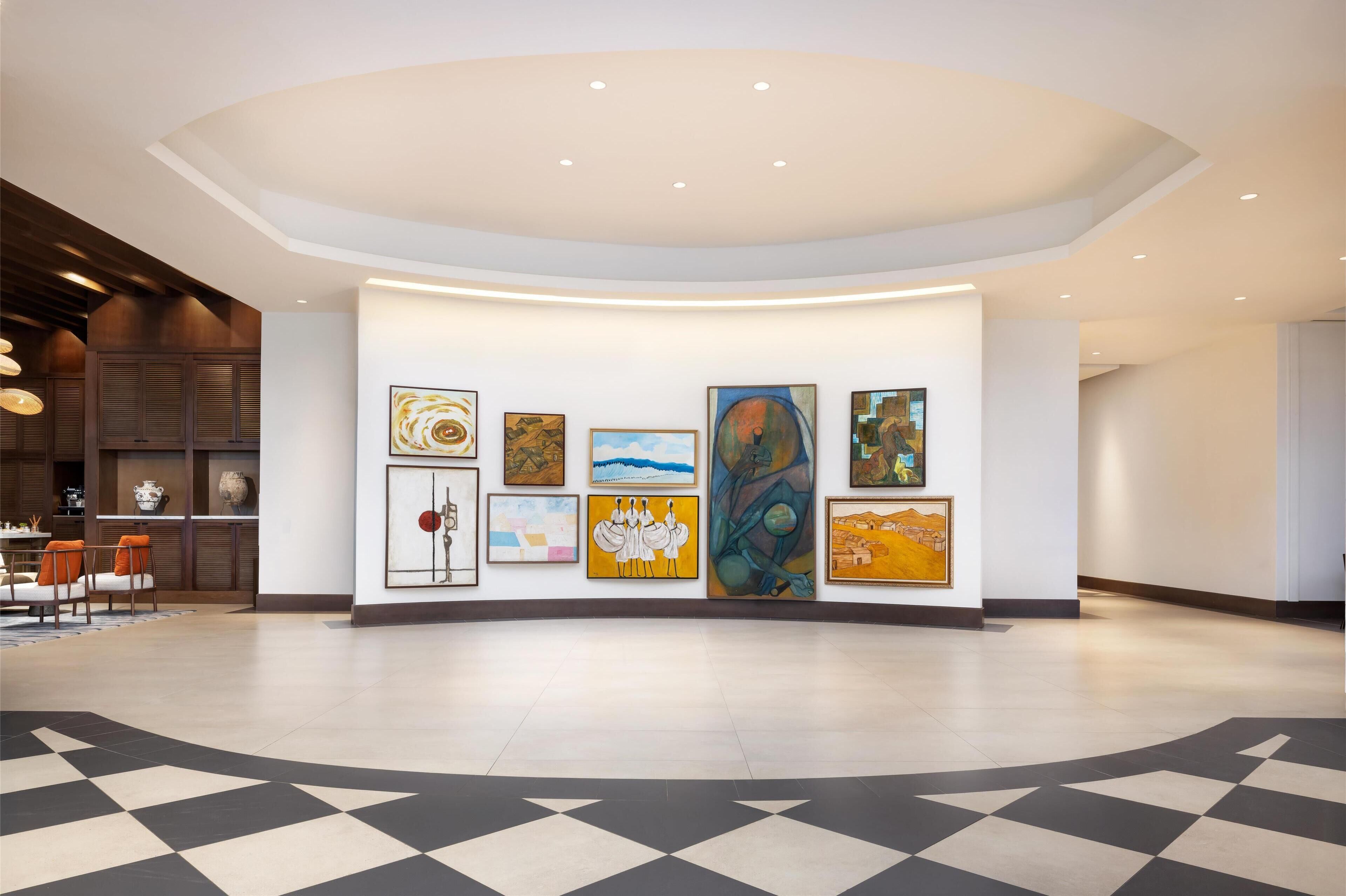 A spacious lobby with a modern design, featuring a large wall of artwork, a checkered floor, and a seating area with orange chairs.