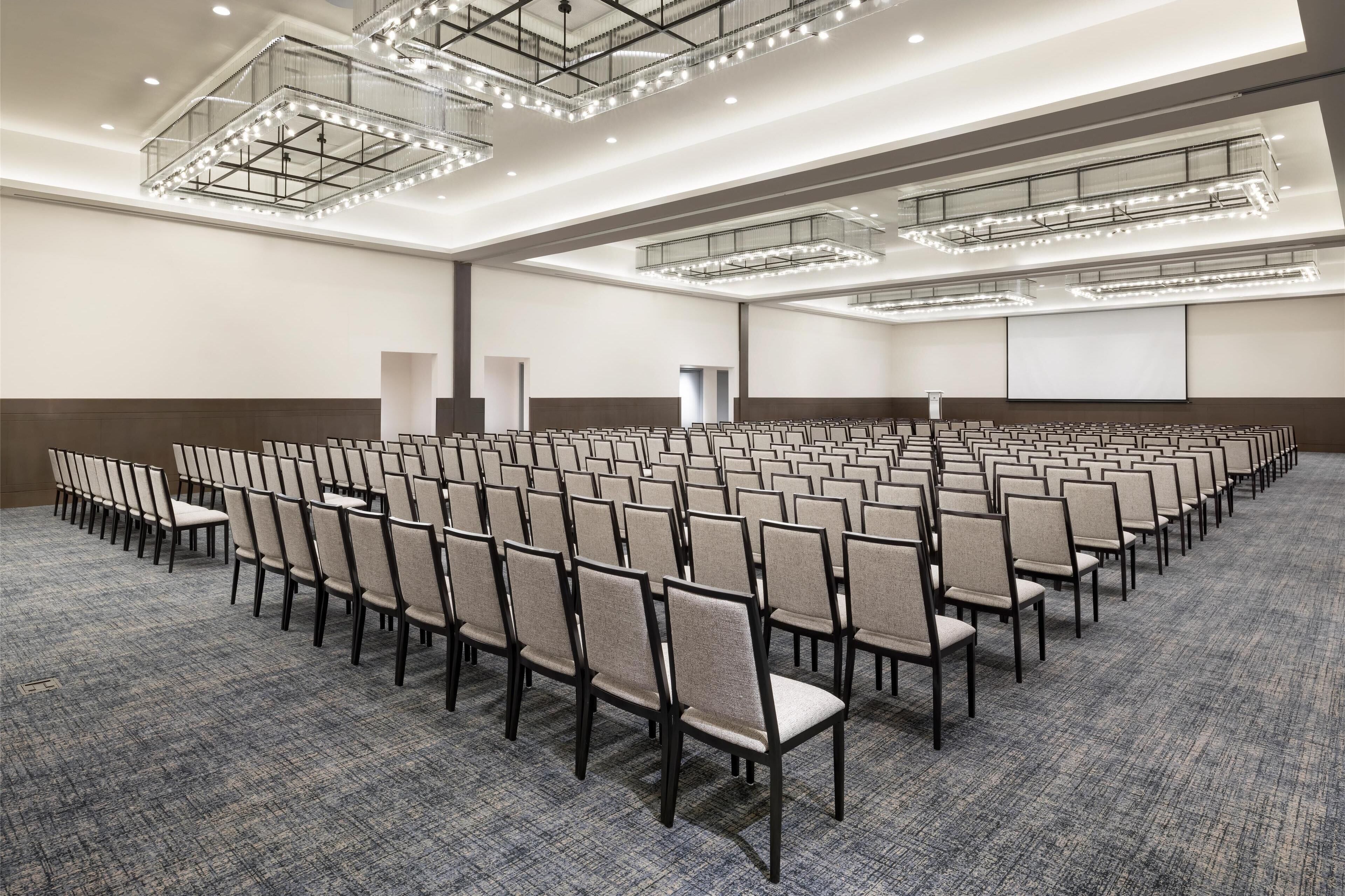 Un amplio salón de conferencias con filas de sillas, piso alfombrado y un techo alto con luminarias.