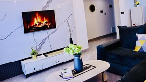 A modern living room with a flat-screen TV mounted on the wall, a coffee table with books and a vase of flowers, a dark blue sofa, and a white entertainment unit with a plant. - spacious 3 bedroom in kilimani gym&pool (Nairobi)