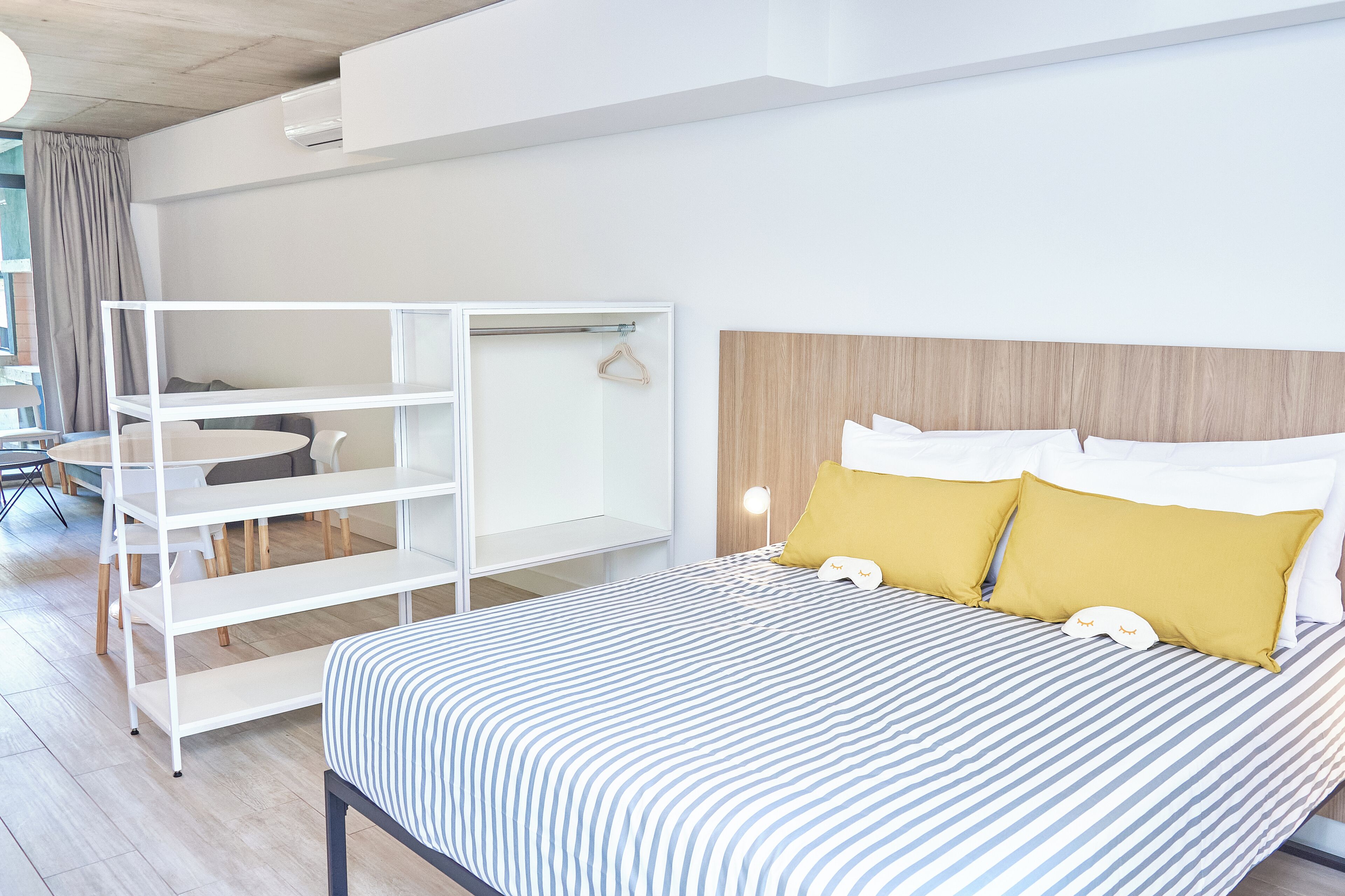Moderni hotellihuone, jossa on sänky, valkoinen hyllykkö, pieni pöytä ja tuoli.