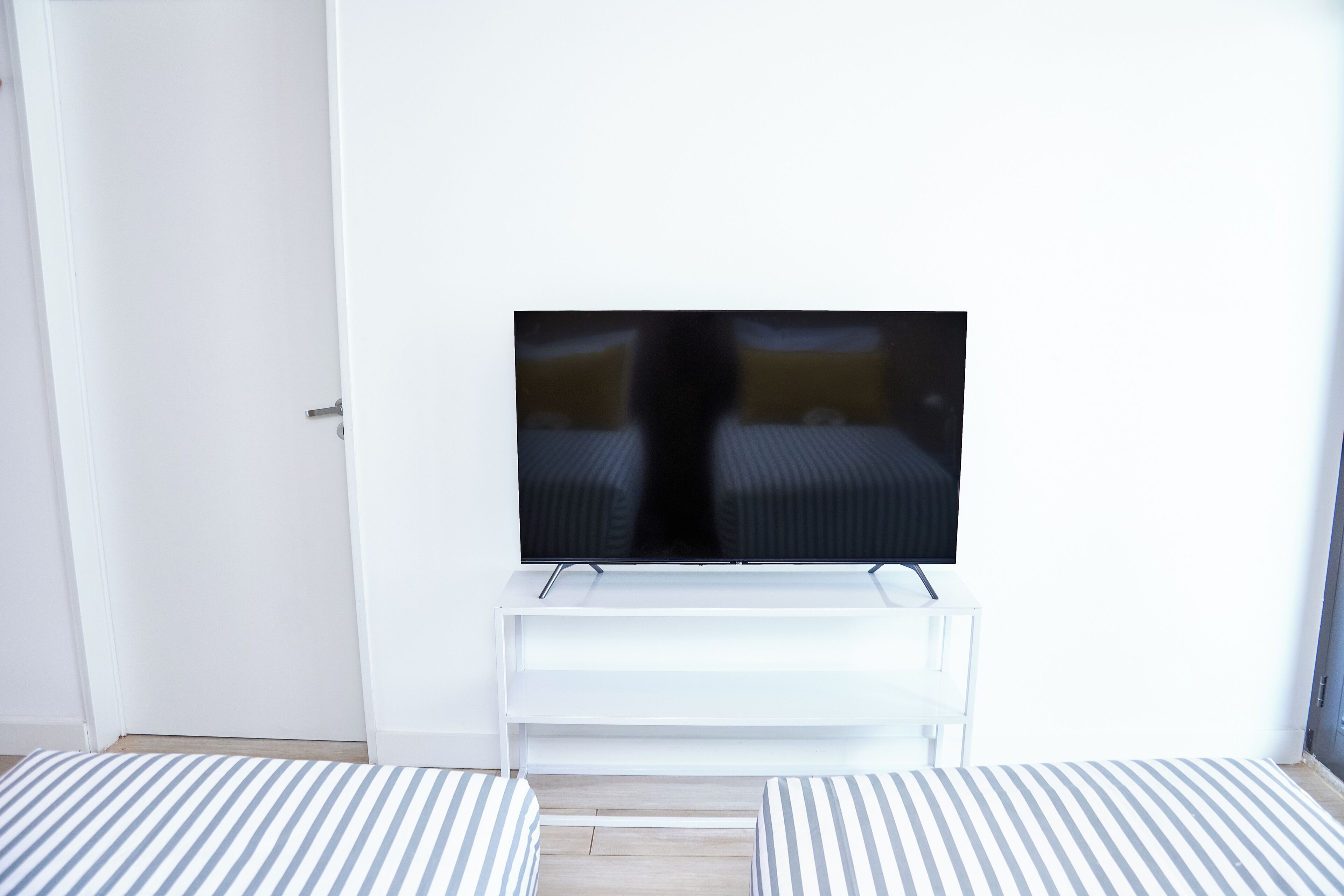 A room with a television on a white stand, a door, and two striped beds.