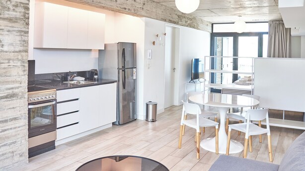 A modern kitchen with white cabinets, a stainless steel refrigerator, and a built-in oven. There is a dining area with a round table and white chairs. - BabelUP (Buenos Aires)