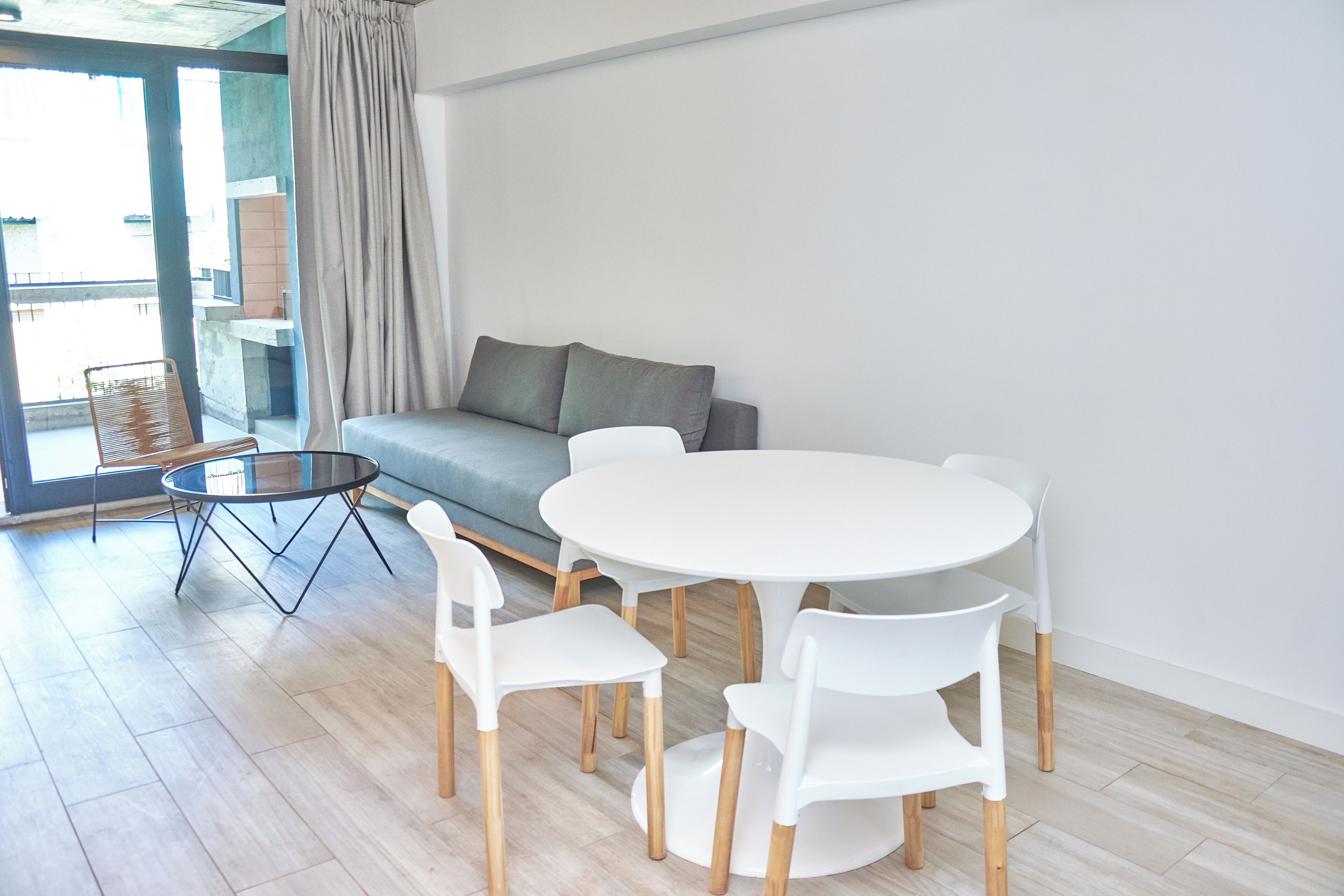 Una sala de estar moderna con una mesa redonda blanca, cuatro sillas blancas, un sofá gris y una silla de mimbre.