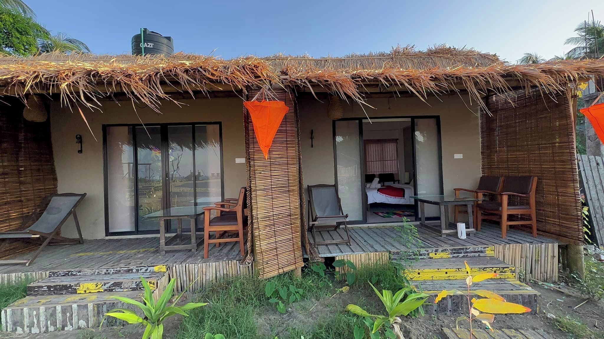 En bungalow med stråtækt tag, en glasdør og et lille udendørs siddeområde med stole og et bord.