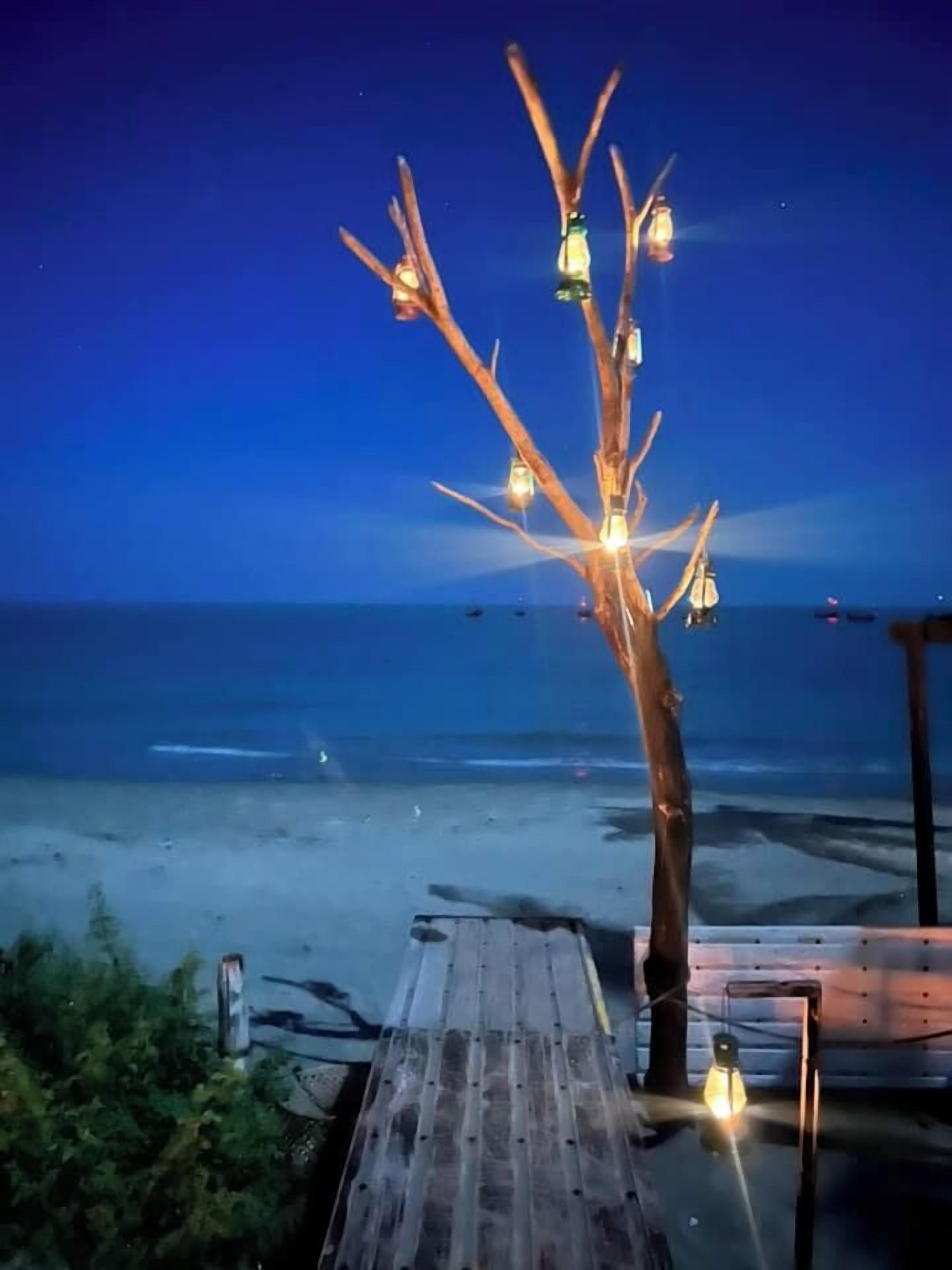 Une terrasse en bois, avec un arbre décoré de lanternes suspendues, donnant sur une plage la nuit.