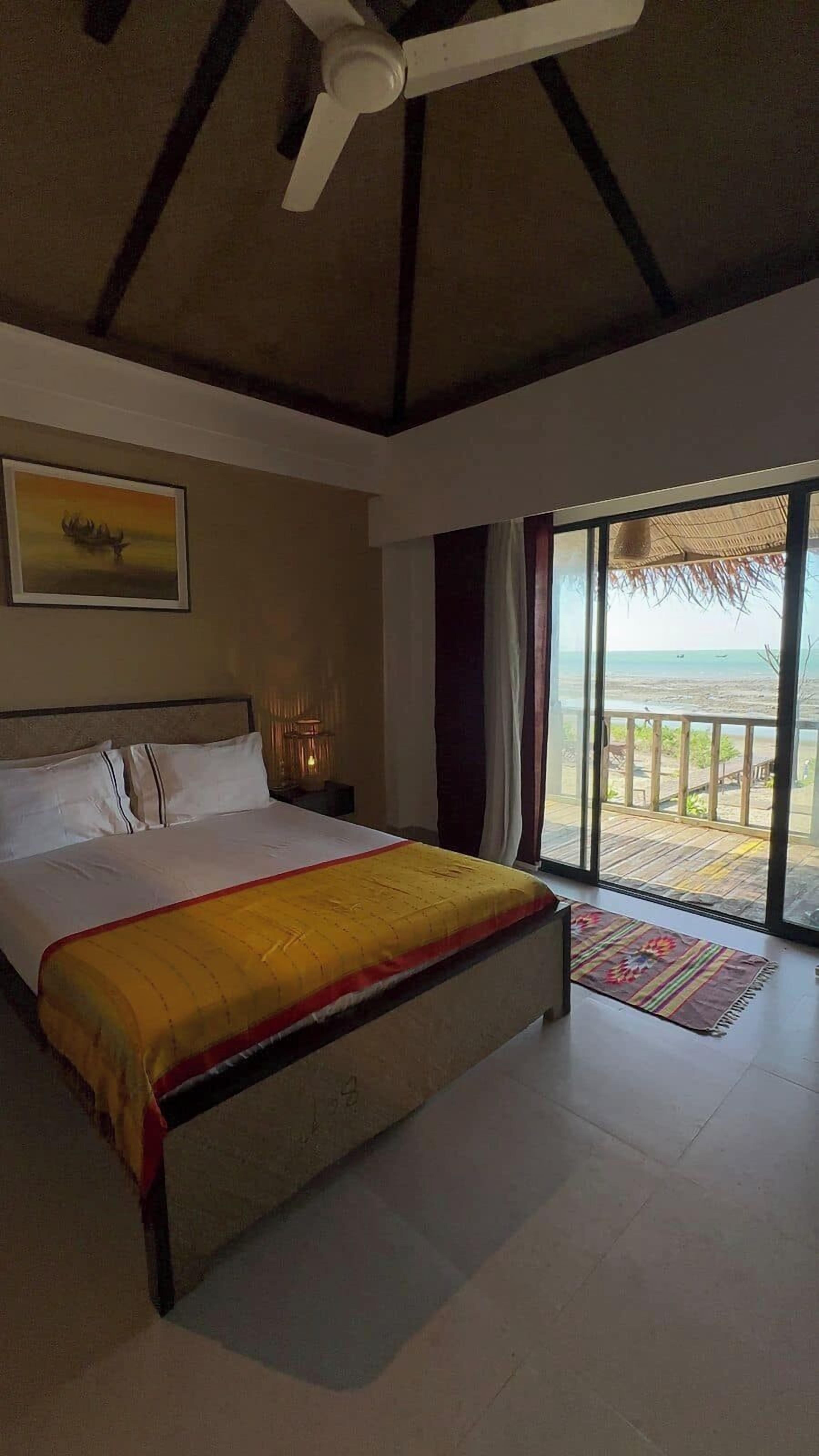 Une chambre avec un lit, un ventilateur de plafond, un tableau et une vue sur la plage.