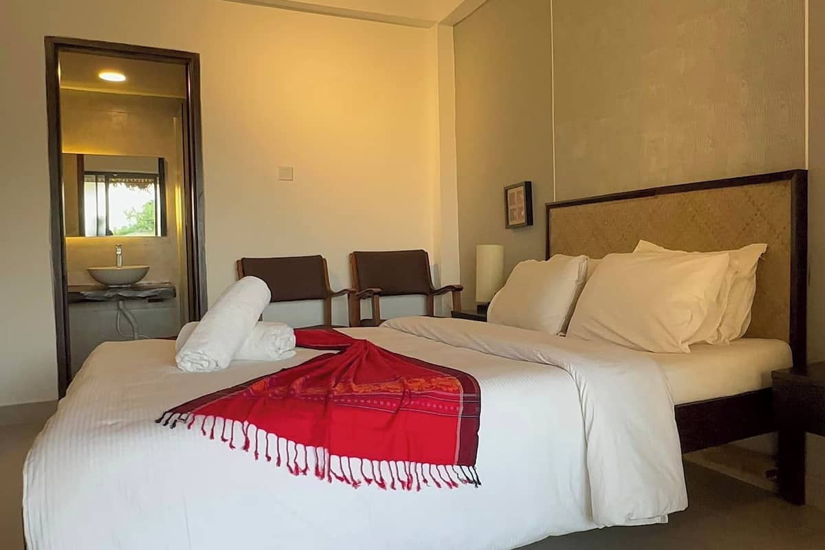 Ein Hotelzimmer mit einem großen Bett, einem Nachttisch mit Lampe, einem Sessel, einem kleinen Tisch mit Schale und einem Spiegel.